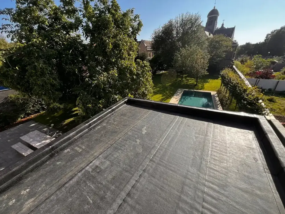 Toiture plate neuve à membrane bitumineuse noire et ses relevés, au-dessus d'un jardin avec piscine et d'un clocher d'église au fond