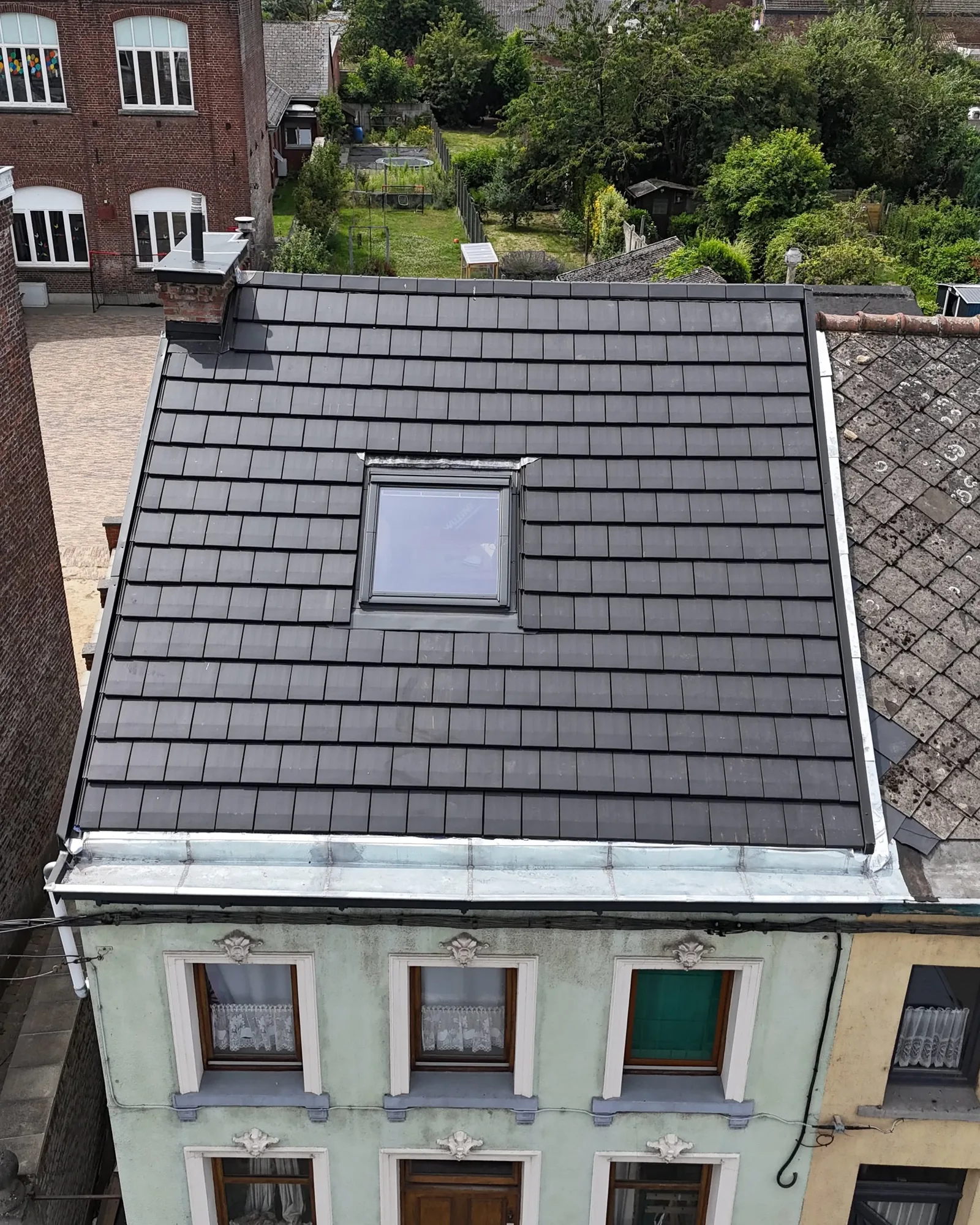 Vue de drone d'un versant de tuiles noires neuves avec fenêtre de toit et solin en zinc, jardins en arrière-plan