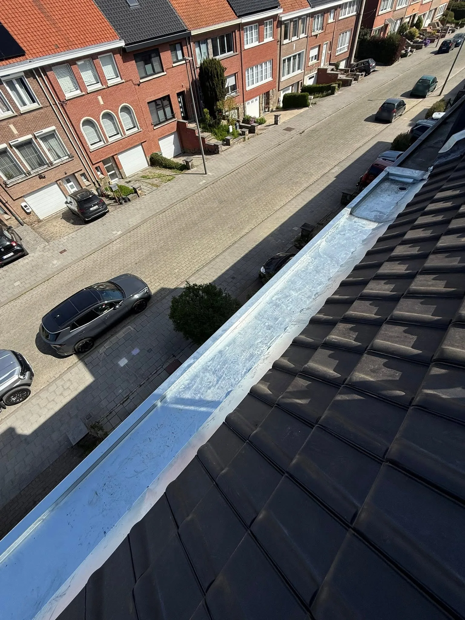 Corniche en zinc naturel posée le long d'un versant de tuiles noires, rue et maisons en briques en contrebas